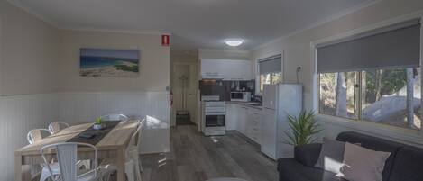 A modern kitchen with white cabinets, a dining area with a wooden table and white chairs, and a grey sofa with a window view.
