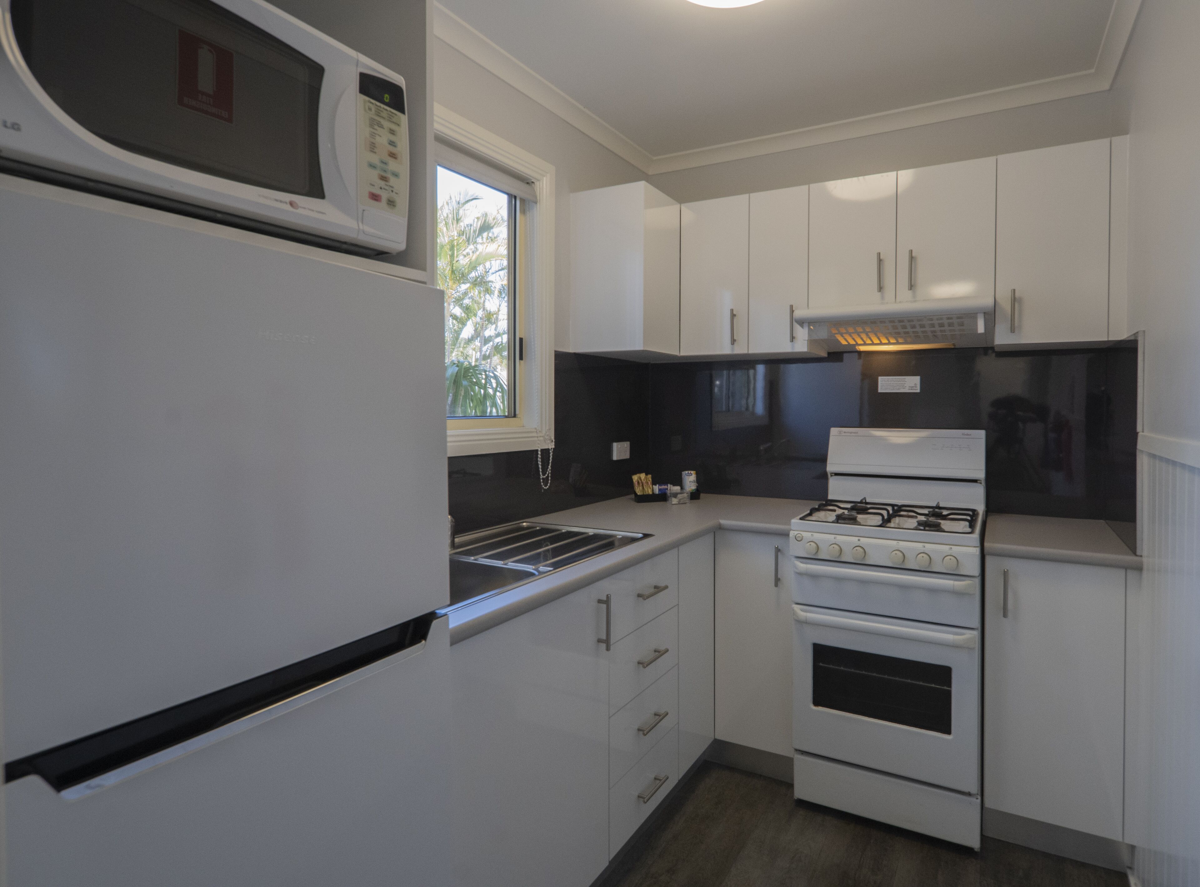 Cocina compacta con armarios blancos, microondas, cocina a gas y ventana que proporciona luz natural.