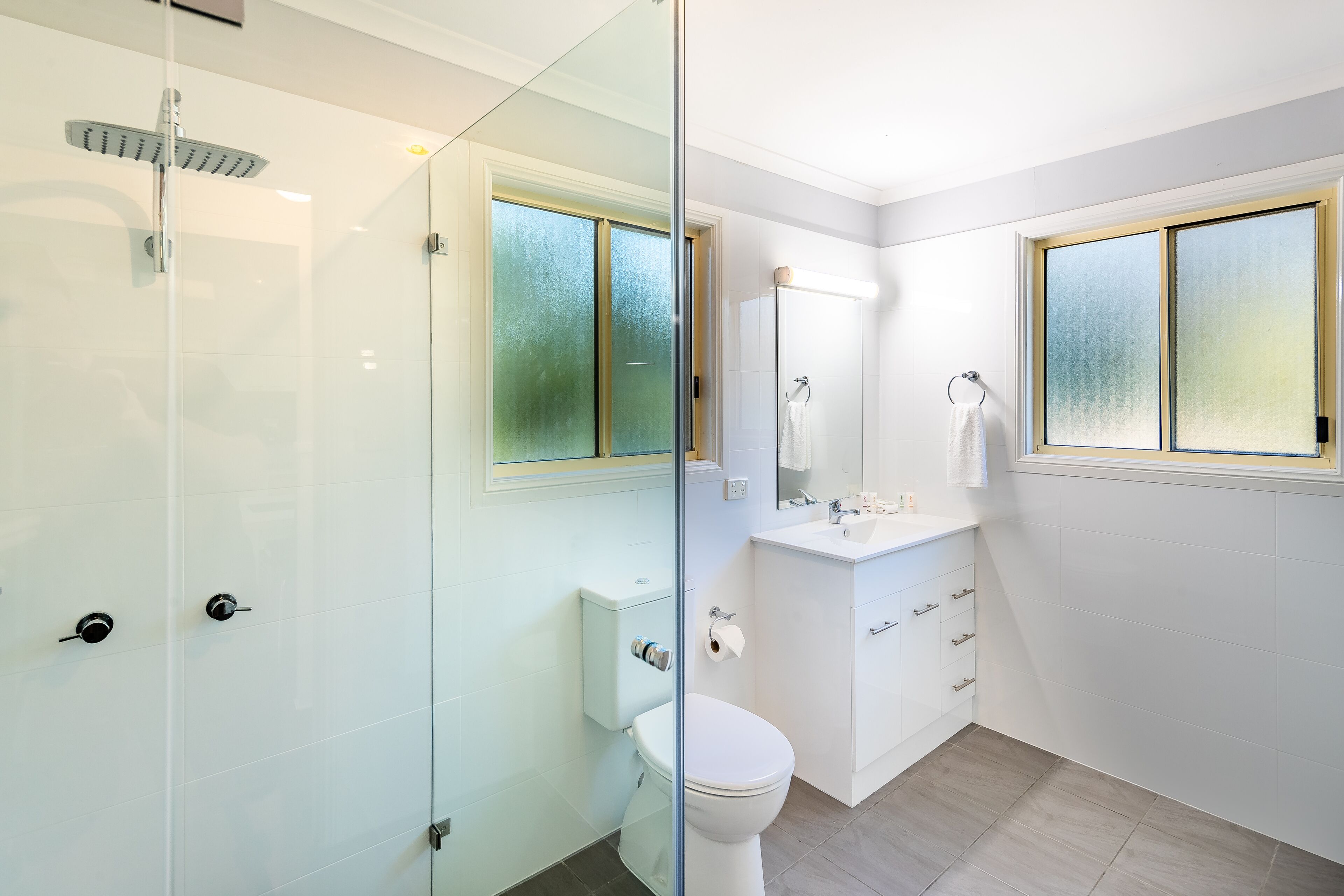 A modern bathroom with a glass shower enclosure, a toilet, a sink with a mirror, and a window.