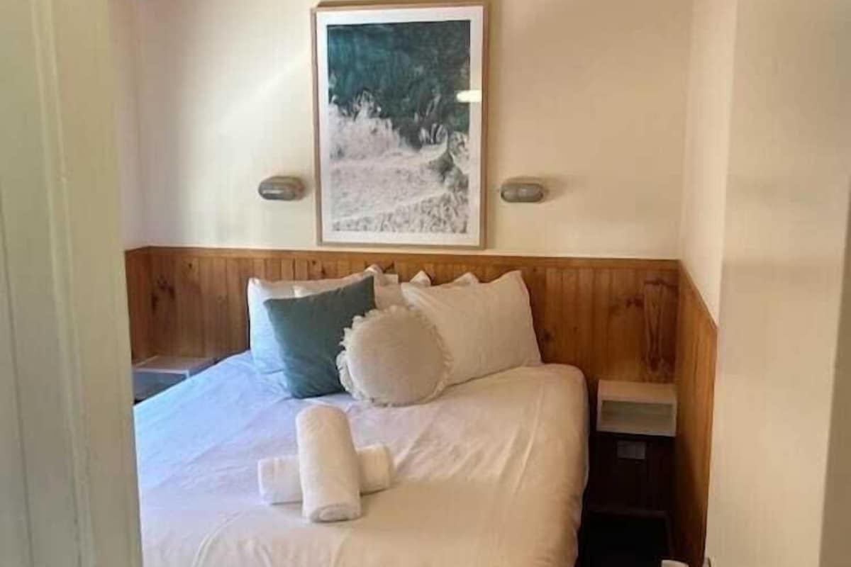 A bedroom with a wooden ceiling, a bed with white bedding, a framed picture on the wall, and a nightstand with a lamp.