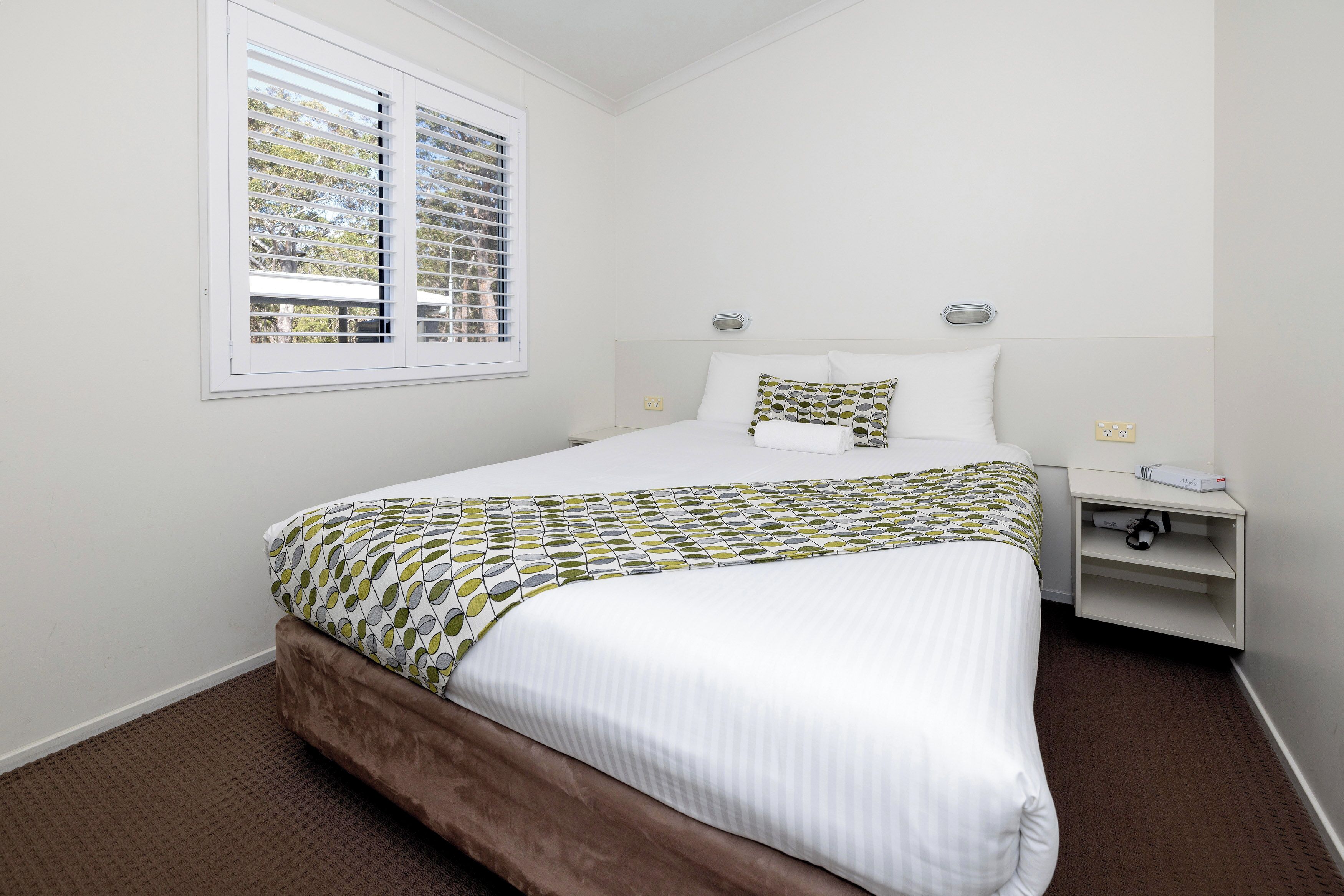 A neatly made bed with a patterned bedspread, a bedside table with a lamp, and a window with blinds.
