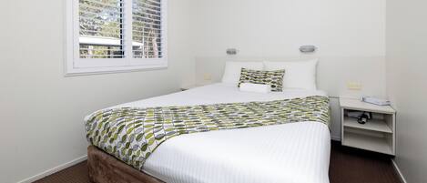 A neatly made bed with a patterned bedspread, a bedside table with a lamp, and a window with blinds.