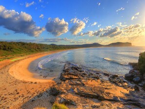 Une plage avec un rivage rocheux au premier plan, une étendue de sable incurvée et, au loin, une chaîne de montagnes sous un ciel partiellement nuageux.
