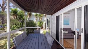 Une terrasse couverte avec une table et des chaises, une porte-fenêtre coulissante donnant sur un salon équipé d’un canapé et orné d’un tableau.