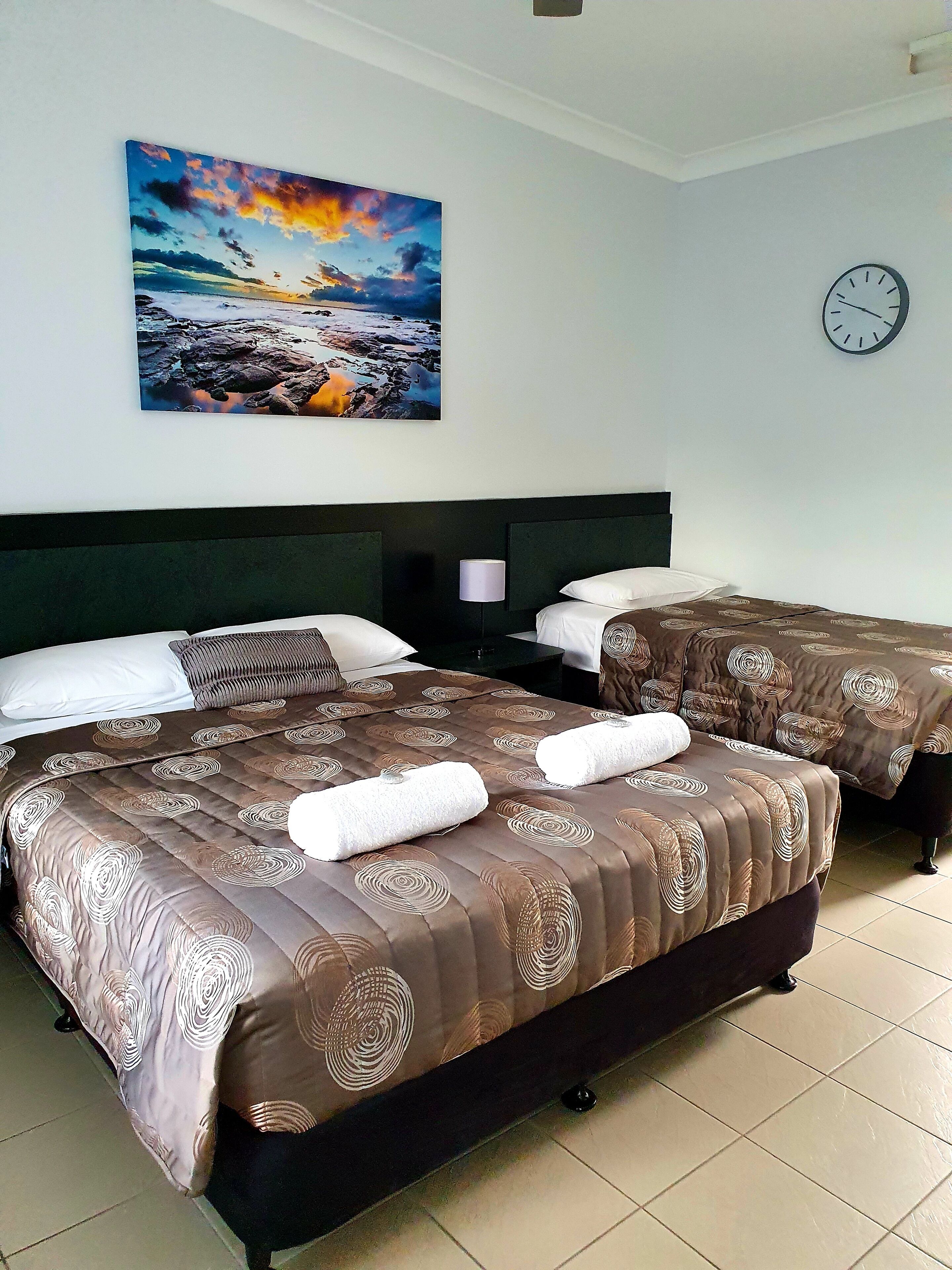 Habitación de hotel con dos camas, un cuadro de una puesta de sol, un reloj en la pared y suelo de baldosas.