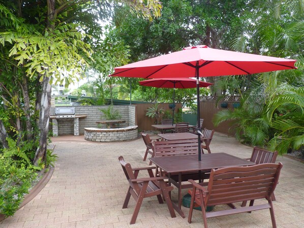 Un patio exterior con suelo de ladrillo, mesas y sillas de madera, un paraguas rojo y una barbacoa de piedra.