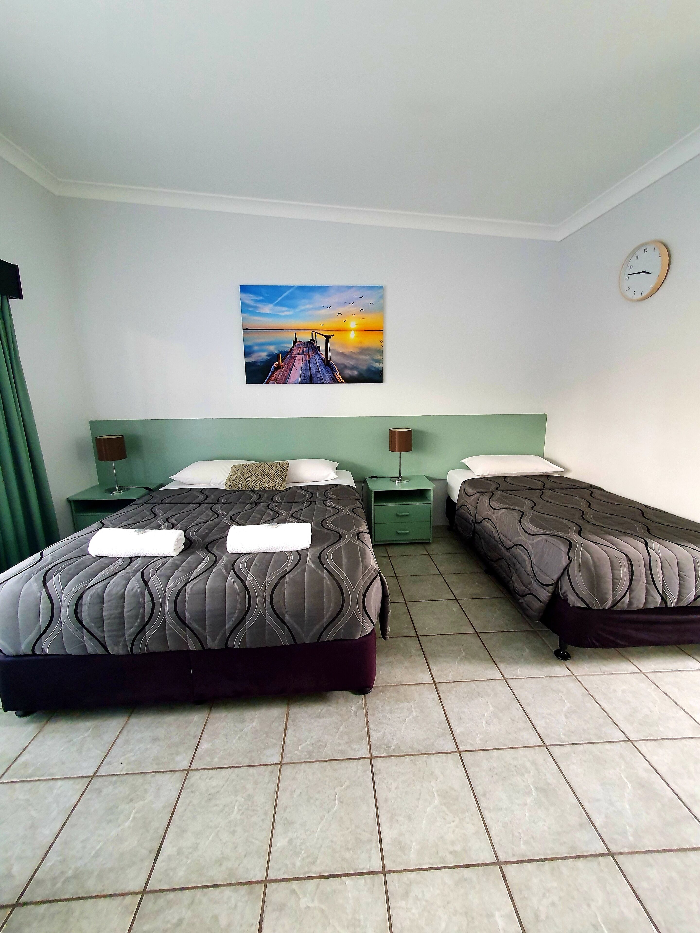 Quarto de hotel com duas camas, um quadro de um pôr do sol, um relógio e cortinas verdes.