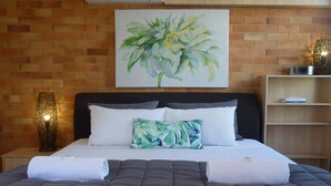 A neatly made bed with a grey quilt, white pillows, and a green leaf-patterned pillow. A painting of a plant hangs above the bed. - Sunseeker Motel (Scarness)