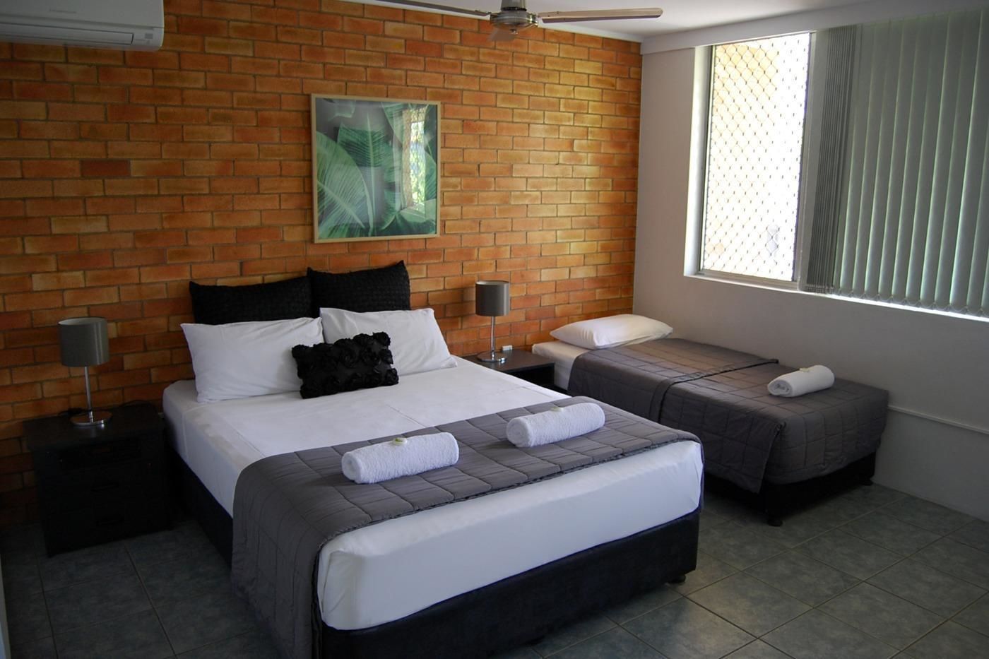 A hotel room with a brick wall, a ceiling fan, a large window with blinds, two beds with white and gray bedding, and a painting on the wall.