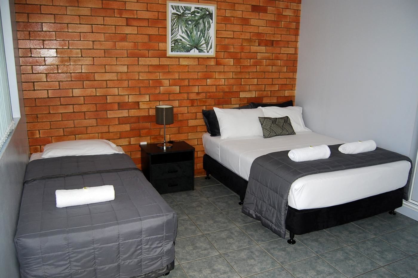 A hotel room with two beds, a brick wall, a framed plant, and a nightstand with a lamp.