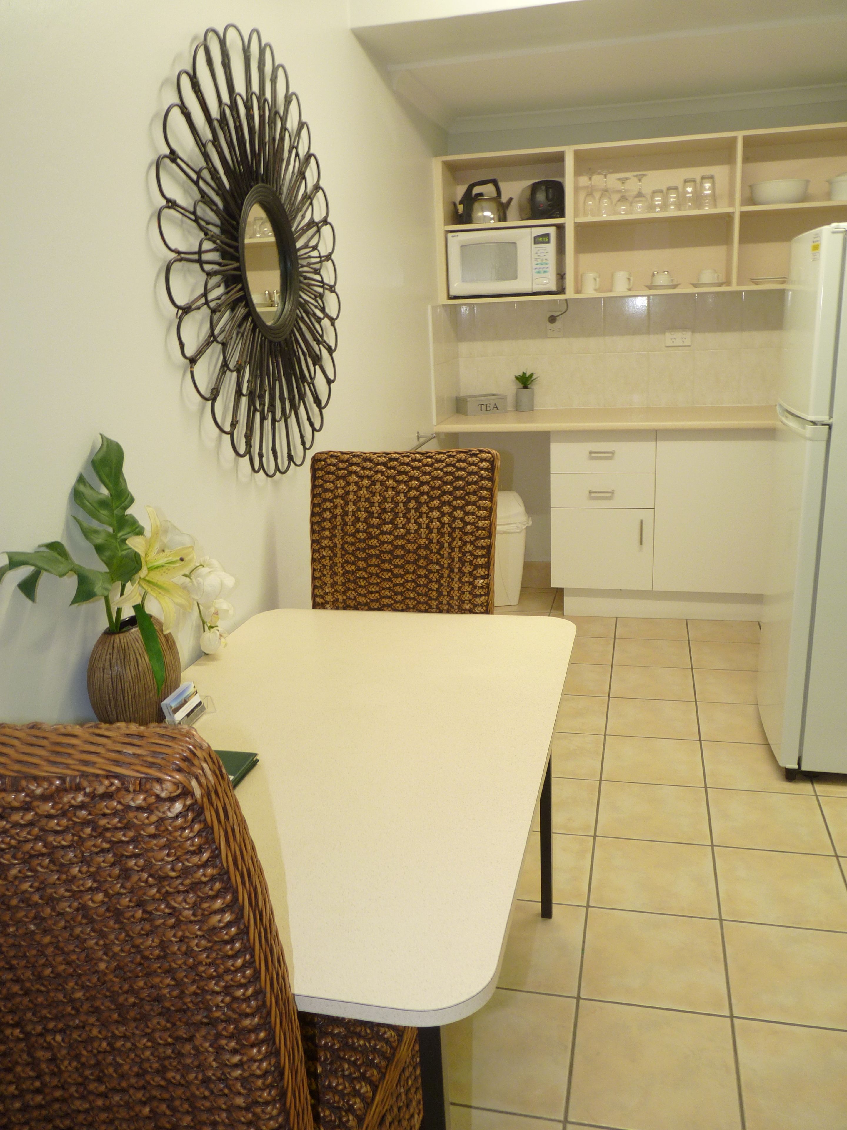 Een eetgedeelte met een tafel en stoelen, een decoratieve spiegel, een vaas met bloemen, en een keuken op de achtergrond met planken en apparatuur.