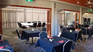 Uma sala de conferências com várias mesas e cadeiras, um palco e um armário de madeira com utensílios de cozinha.