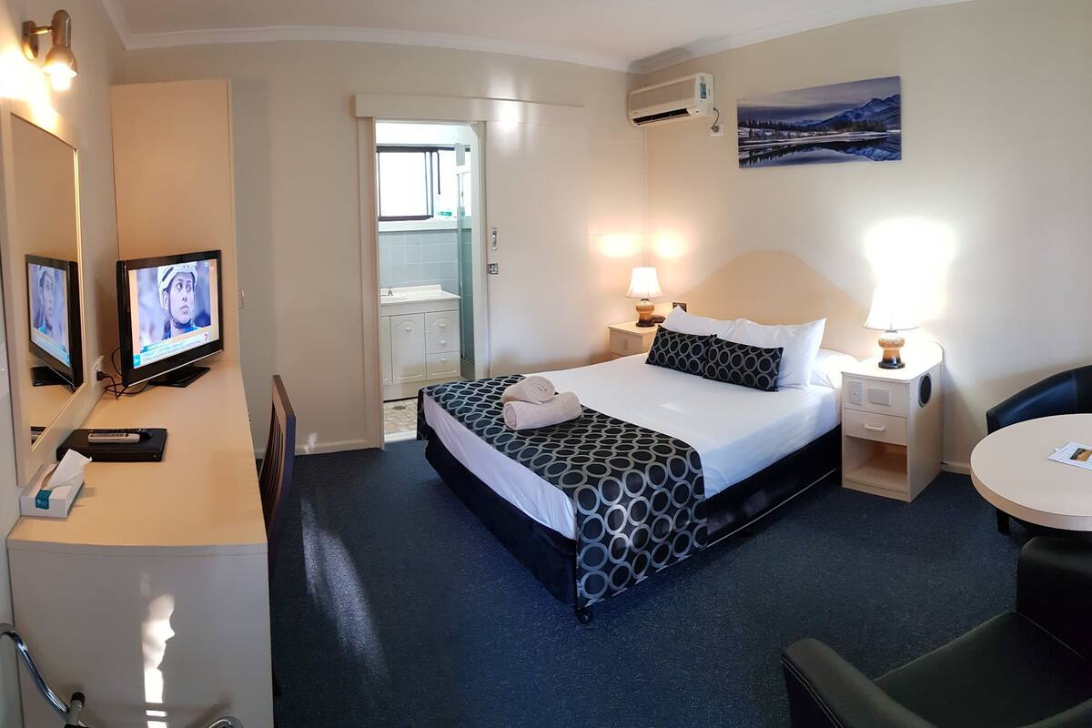 A hotel room with a bed, bedside tables, a desk, a chair, a TV, and a painting on the wall.