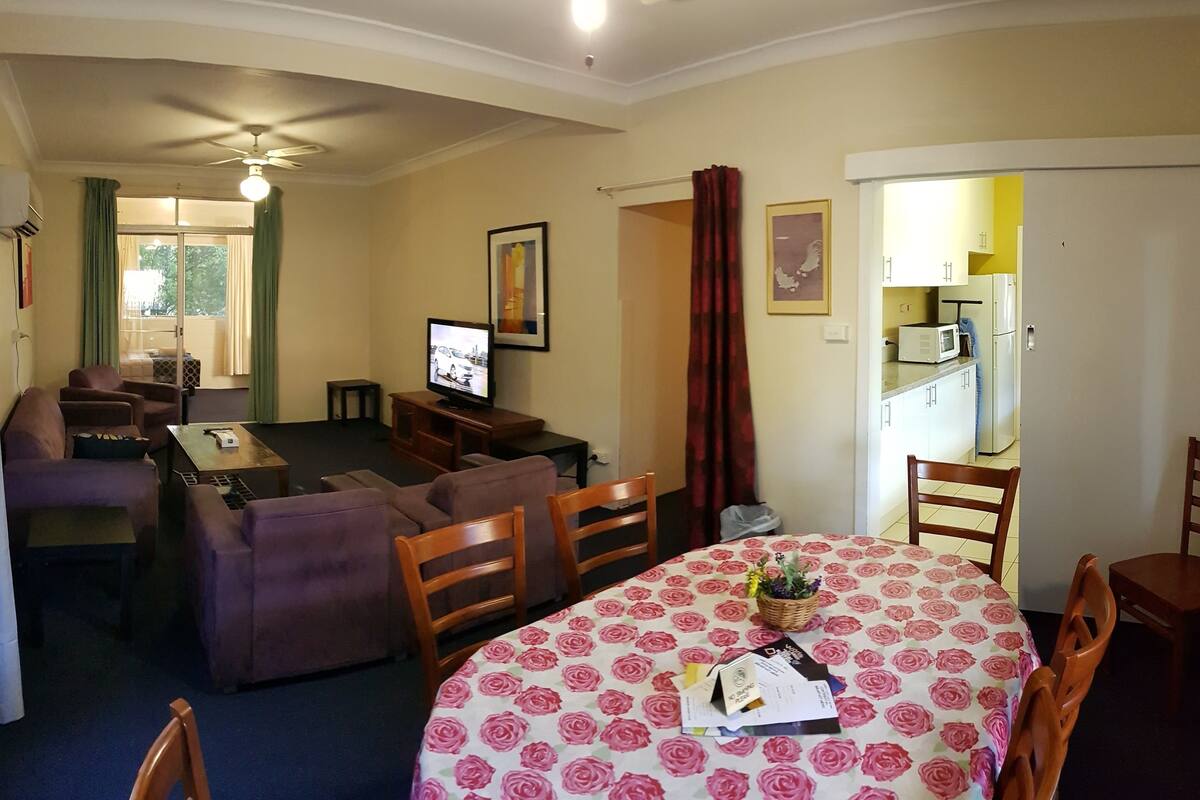 A living room with a television, sofa, and dining table with chairs. There is a kitchen area visible through an open door.