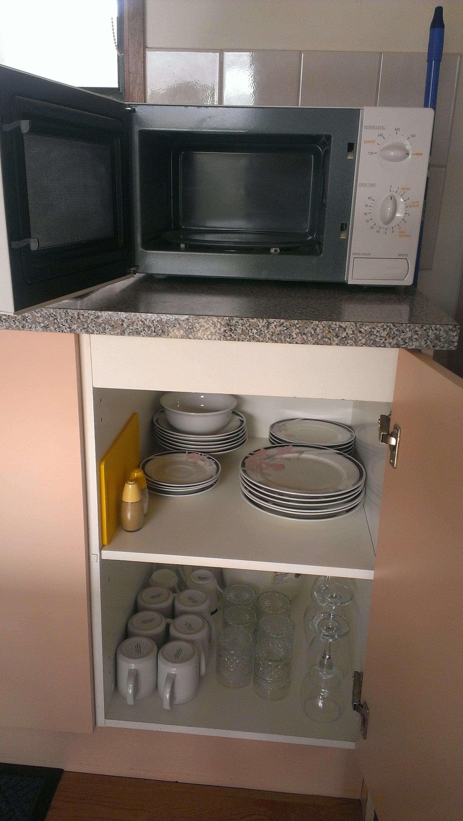 Una cucina con un forno a microonde, un pensile pieno di piatti e uno scaffale con tazze e bicchieri.