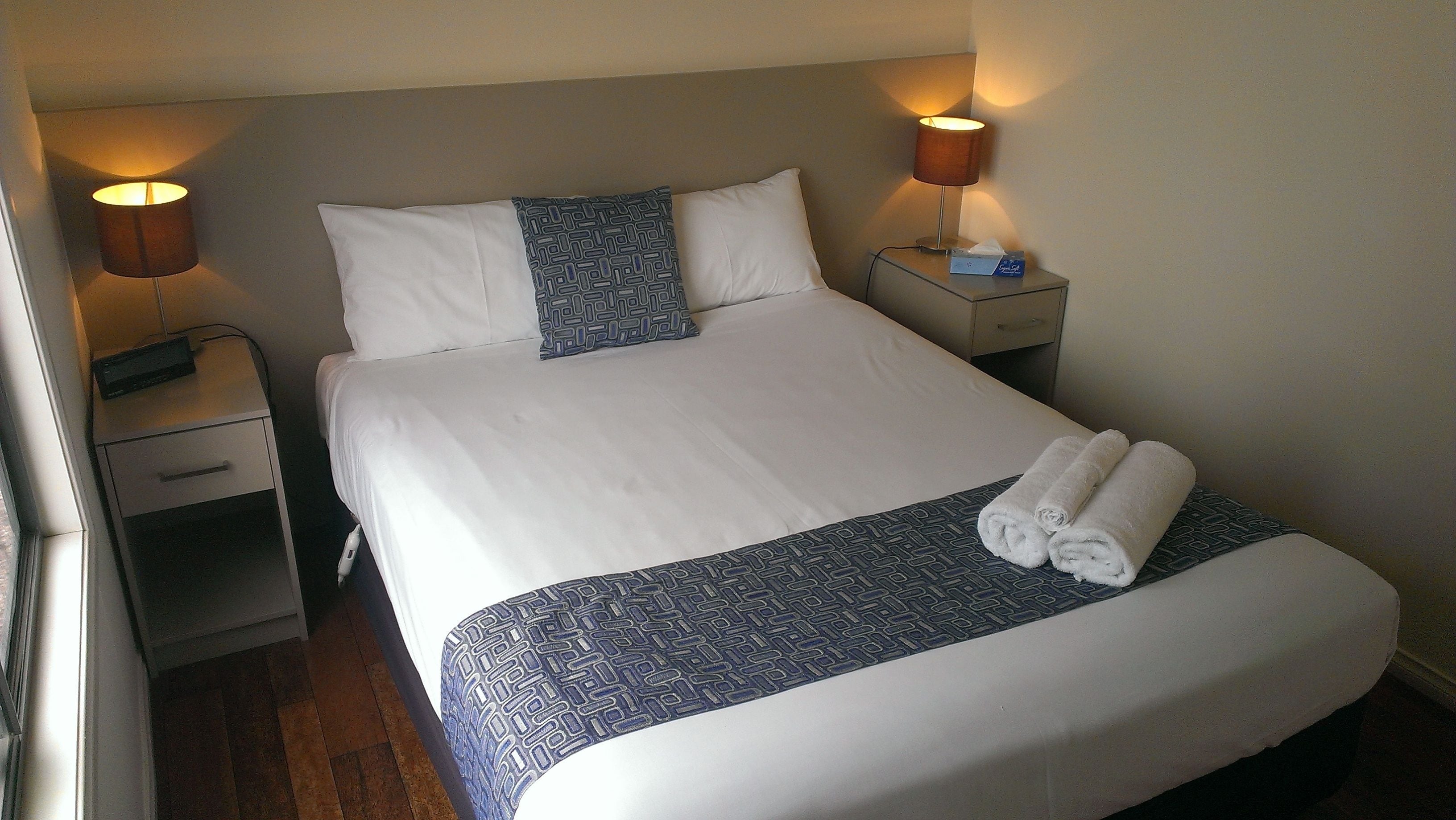 A neatly made bed with white and blue bedding, two bedside lamps, and a small bedside table with a tissue box.