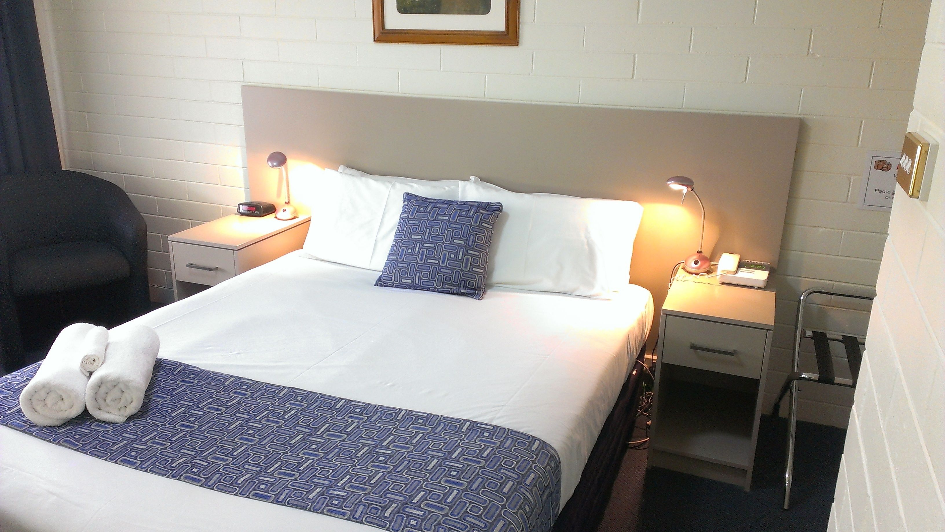 A neatly made bed with a patterned blanket, two bedside lamps, a bedside table with a phone, and a chair in a hotel room.
