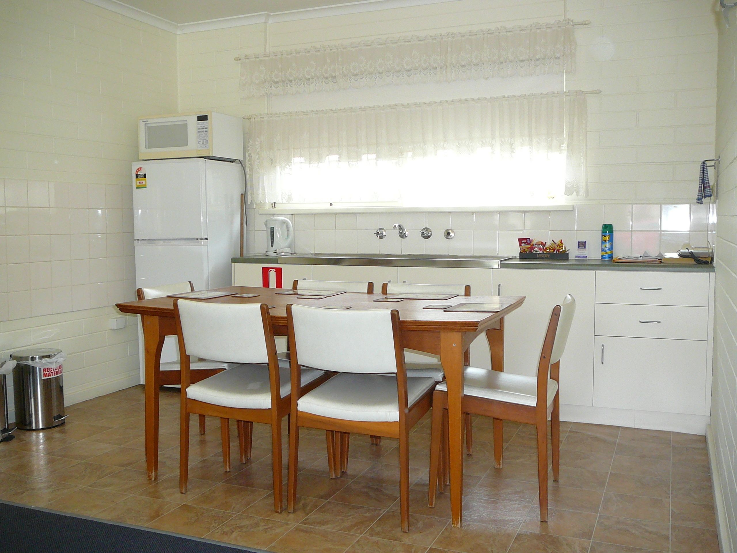 Een compacte keuken met witte apparaten, een eettafel met stoelen en een raam met dunne gordijnen.