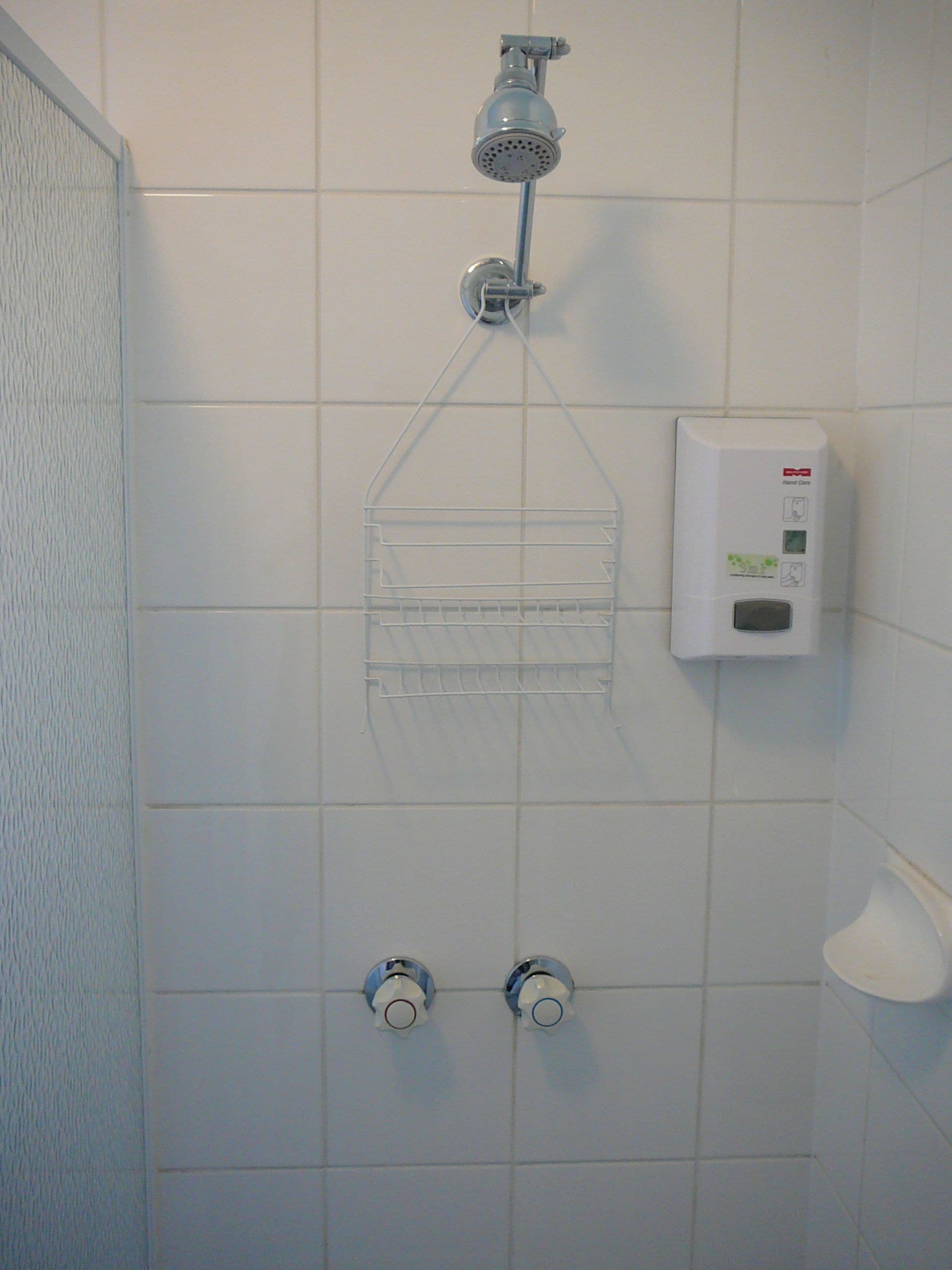 A shower with a handheld showerhead, a soap dispenser, and a clear plastic shelf.