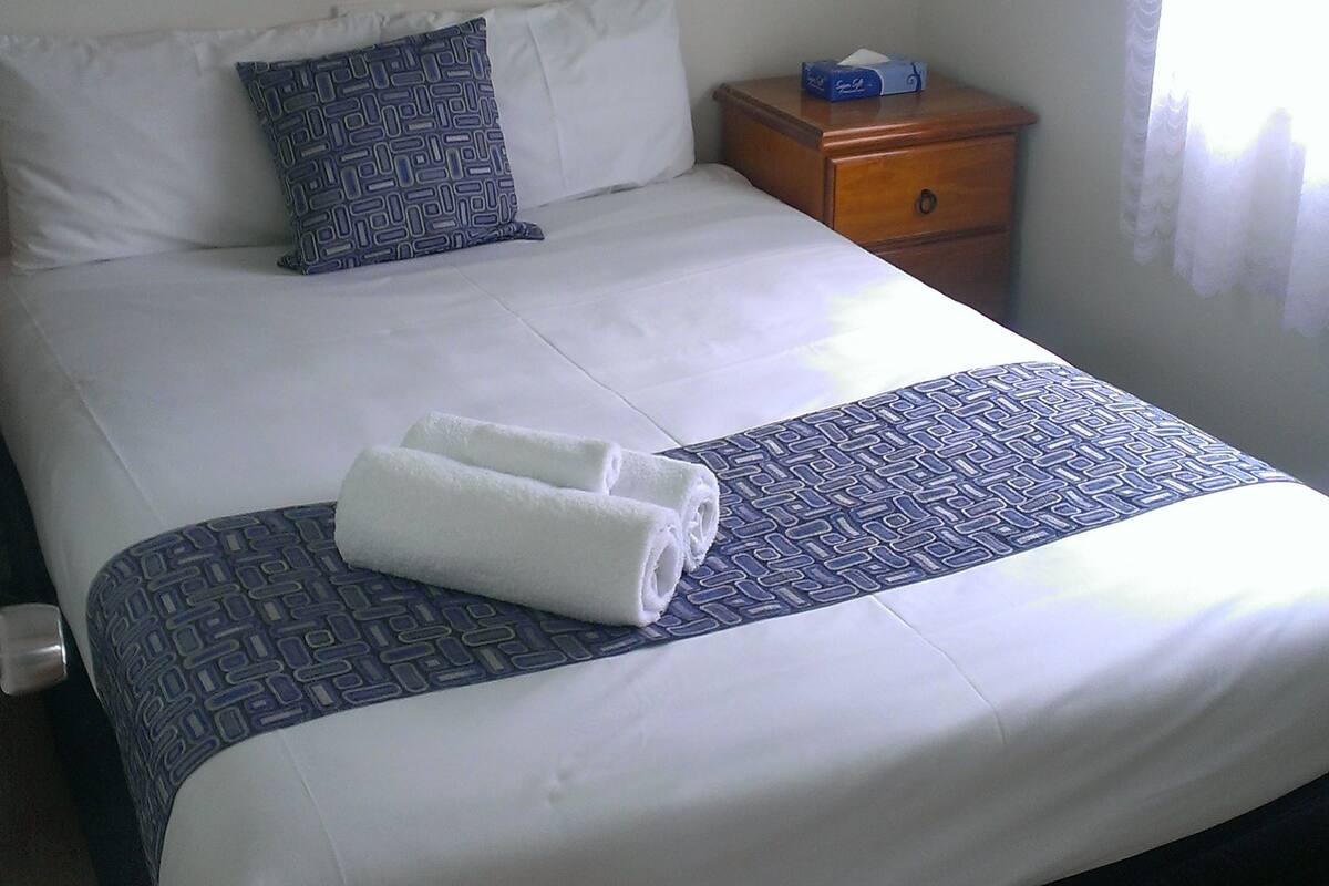 Ein ordentlich bezogenes Bett mit einem gemusterten Überwurf, einem Kopfkissen und einem zusammengerollten Handtuch. Ein kleiner Nachttisch mit Taschentuchbox und einem gerahmten Bild befindet sich darüber.