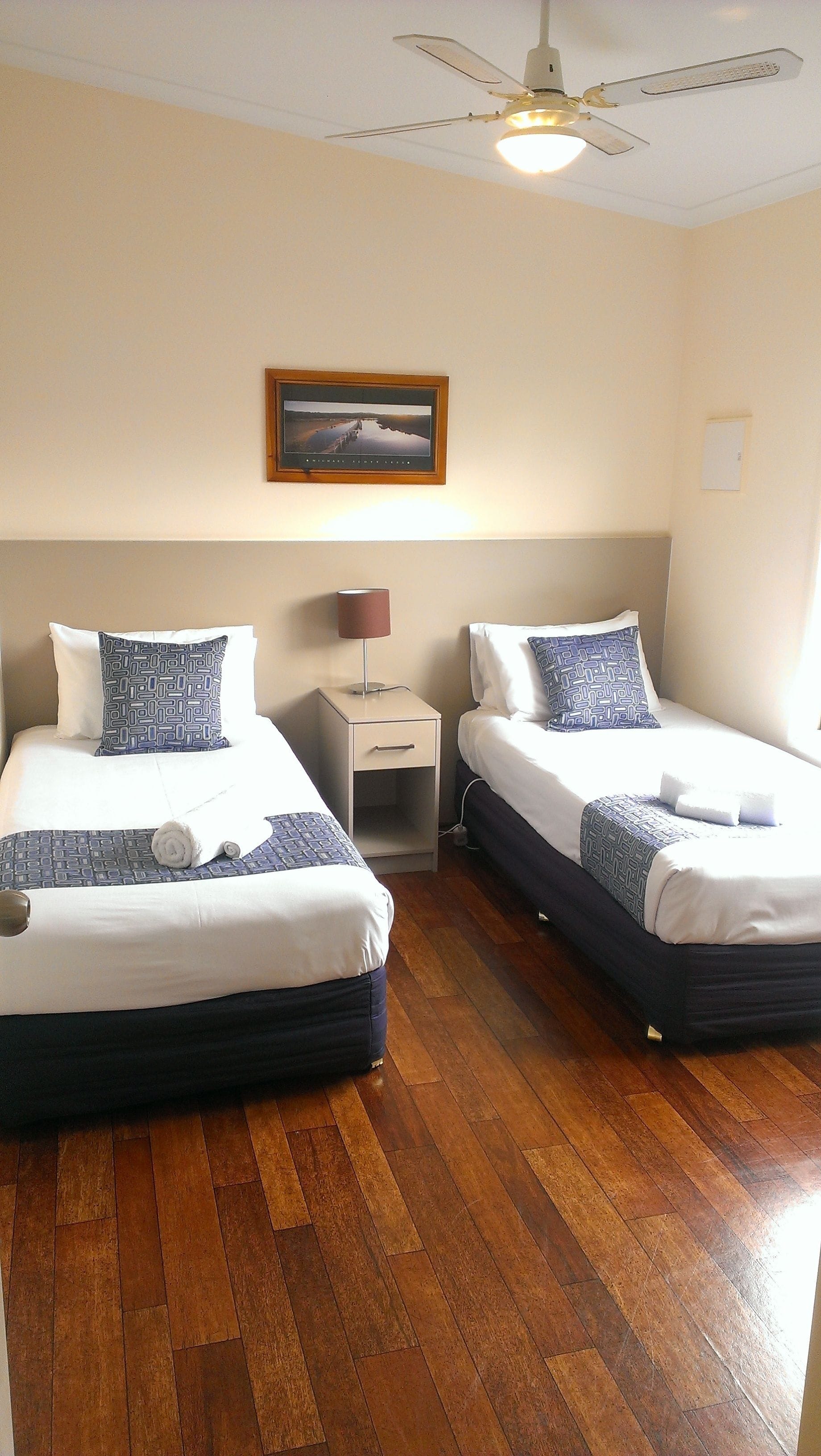 A hotel room with two beds, wooden flooring, a ceiling fan, a nightstand, and a framed picture on the wall.