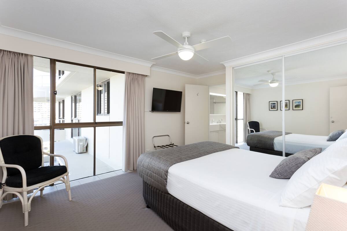 Une chambre d’hôtel avec un grand lit, une chaise, un ventilateur de plafond, une télévision et une porte-fenêtre coulissante donnant sur un balcon.