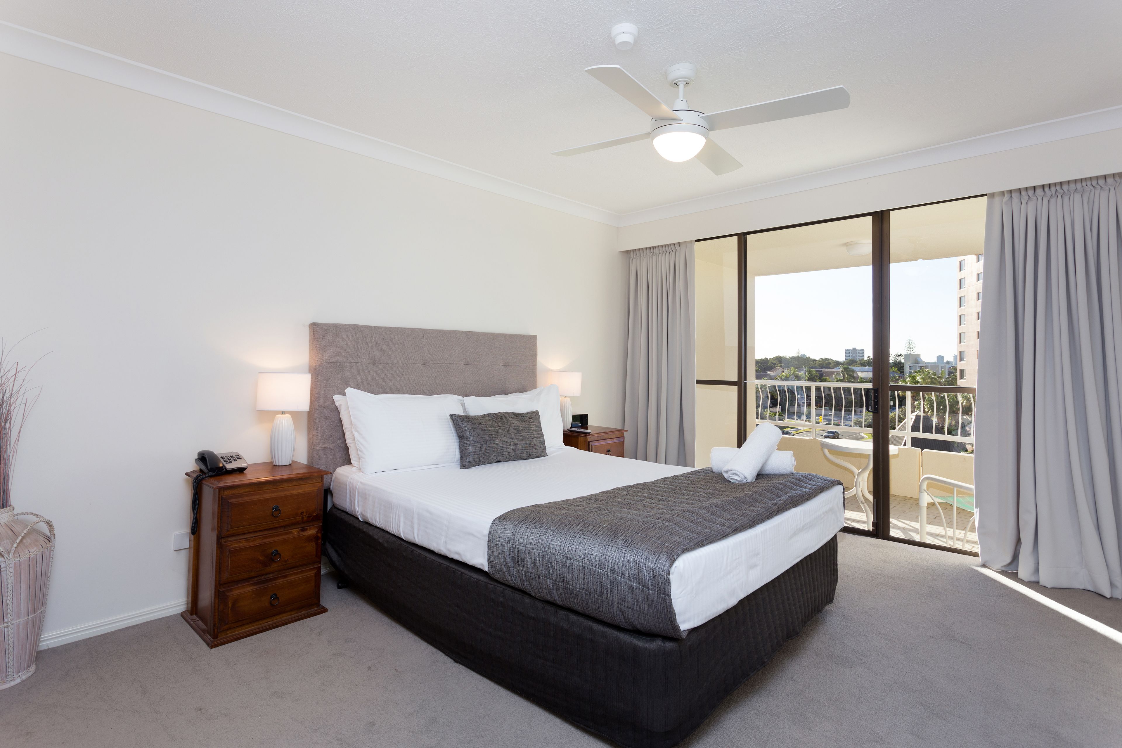 Une chambre d’hôtel avec un grand lit, une table de chevet, une lampe, un ventilateur de plafond et un balcon offrant une vue sur la ville.