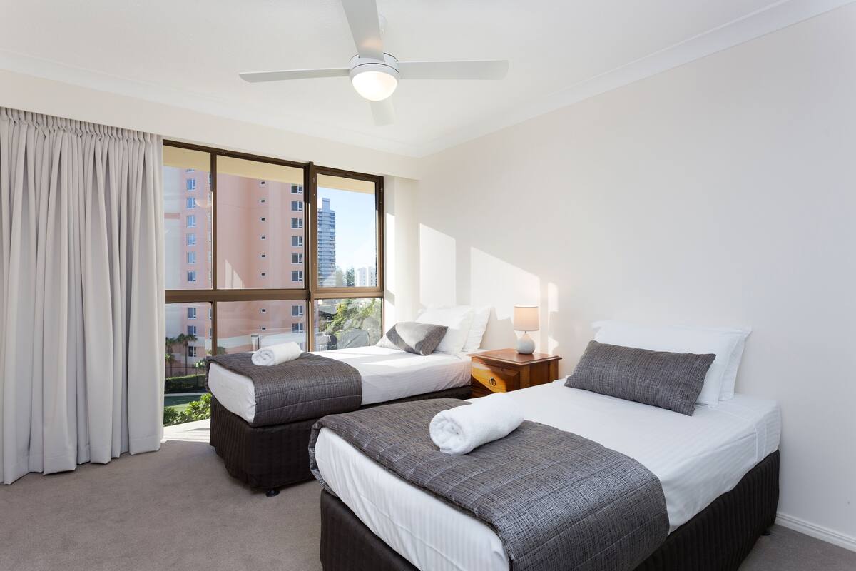 Une chambre d’hôtel avec deux lits, un ventilateur de plafond, une fenêtre avec des rideaux et une vue sur les bâtiments environnants.