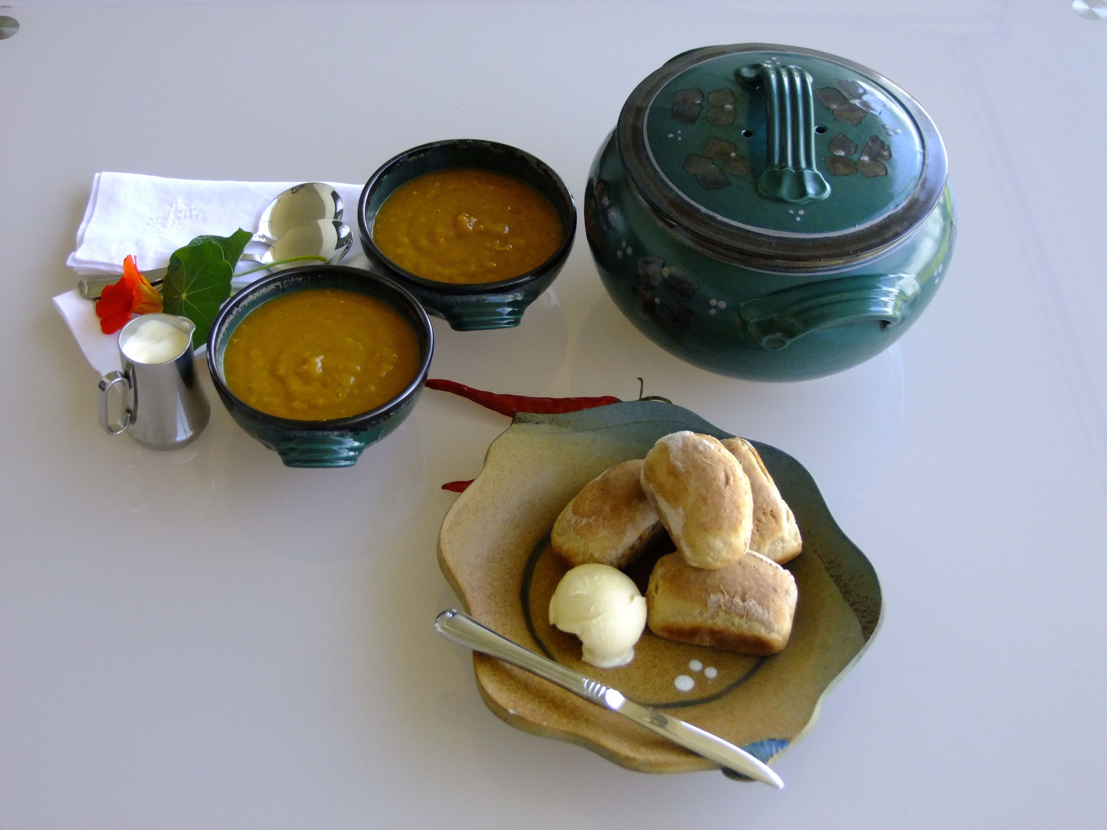 Et bord med tre skåle suppe, en tepotte, et fad med brød og smør, samt en lille kande.