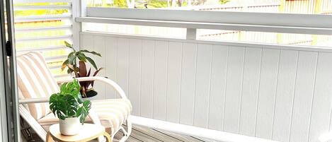 A wooden deck with a white railing, a wicker chair, a small table with a potted plant, and a view of trees and a fence.