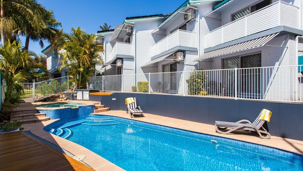 Una piscina con sillas de descanso, una terraza de madera y un edificio blanco con balcones.