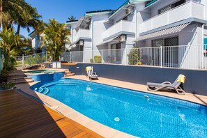 Una piscina con sillas de descanso, una terraza de madera y un edificio blanco con balcones.