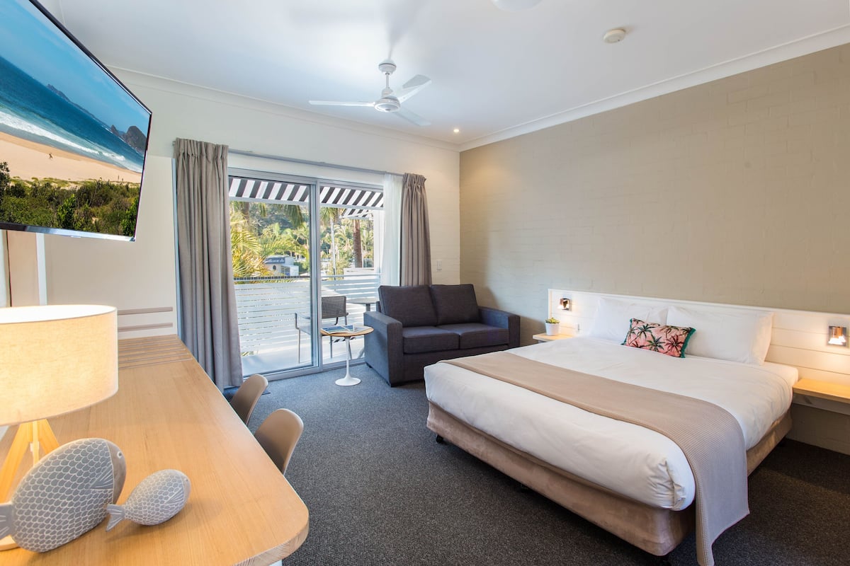 A hotel room with a bed, a sofa, a desk with a lamp, a TV, and a view of the beach through the window.