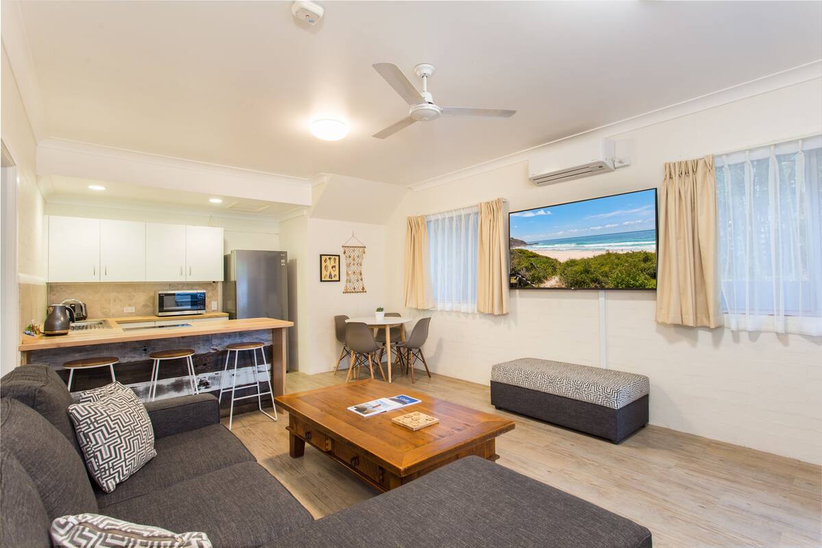 A modern living room with a sofa, a coffee table, and a TV. There is a kitchen area with a microwave and a kettle. The room has a ceiling fan and a window with curtains.