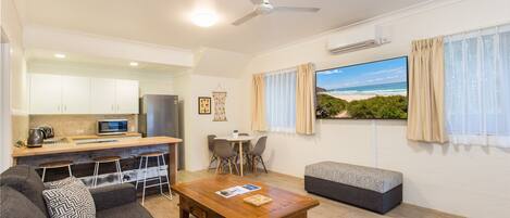A modern living room with a sofa, a coffee table, and a TV. There is a kitchen area with a microwave and a kettle. The room has a ceiling fan and a window with curtains.