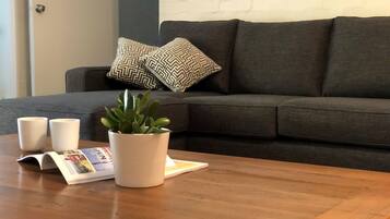 A living room with a dark grey sofa, two patterned cushions, a white potted plant, two white cups, and a magazine on a wooden coffee table.
