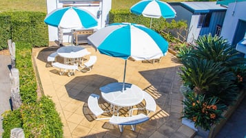 Een terras met buitenmeubilair en parasols, omringd door een haag en een gebouw.