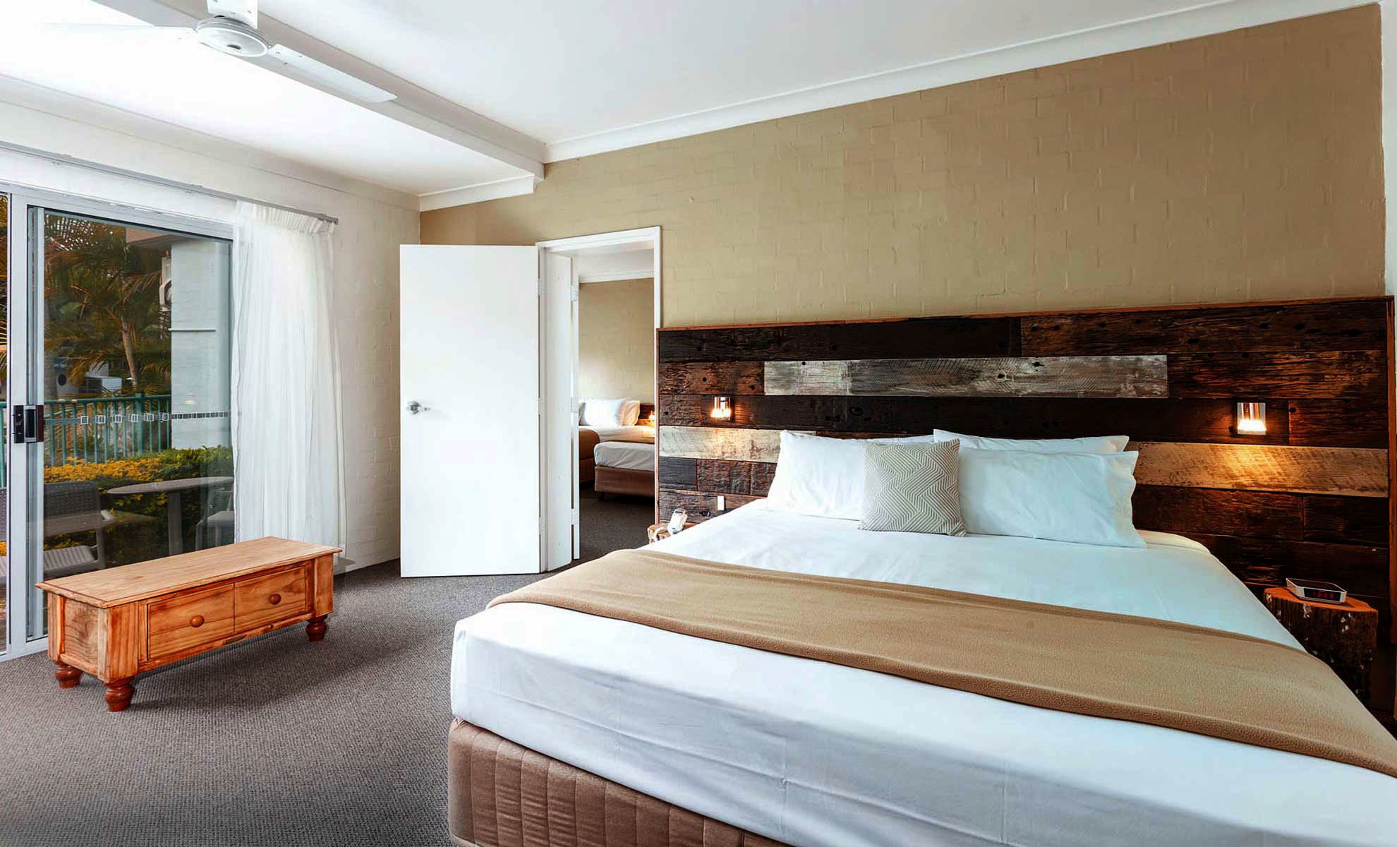 Una habitación de hotel moderna con una cama grande, una mesita de noche de madera, un pequeño aparador de madera y un ventanal corredero que da a un balcón.