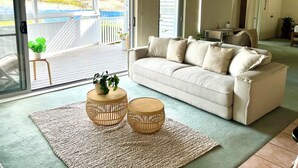 Un salón con un sofá color beige, dos mesitas de madera redondas, un tapete tejido y vista a una piscina a través de un ventanal.