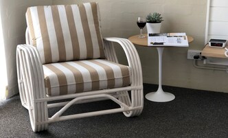 Un sillón a rayas con un cojín, una pequeña mesa redonda con una planta en maceta y una copa de vino, y un reloj digital en una estantería.