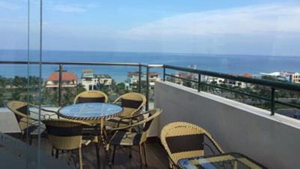 A balcony with wicker furniture and a glass table, overlooking a coastal view with buildings in the distance. - Dattravi Hostel (Da Nang)