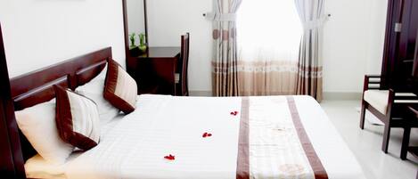 Un lit bien fait, recouvert d’une couvre-lit à rayures blanches et brunes, avec une tête de lit en bois et quelques oreillers. Des pétales de rose sont disposés sur le lit, et on aperçoit une chaise à l’arrière-plan.