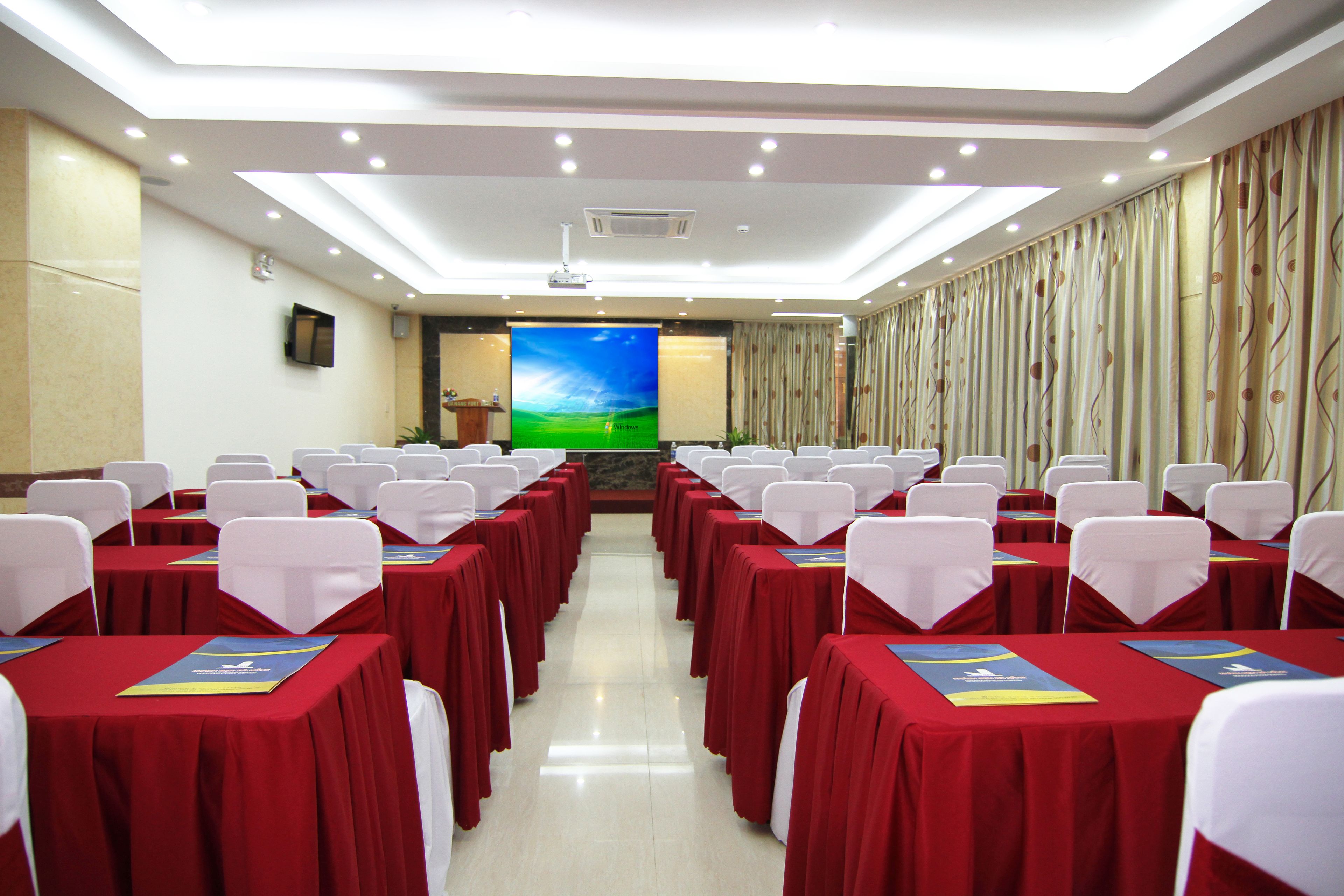 A conference room with rows of tables, white chairs, and a large screen at the front.