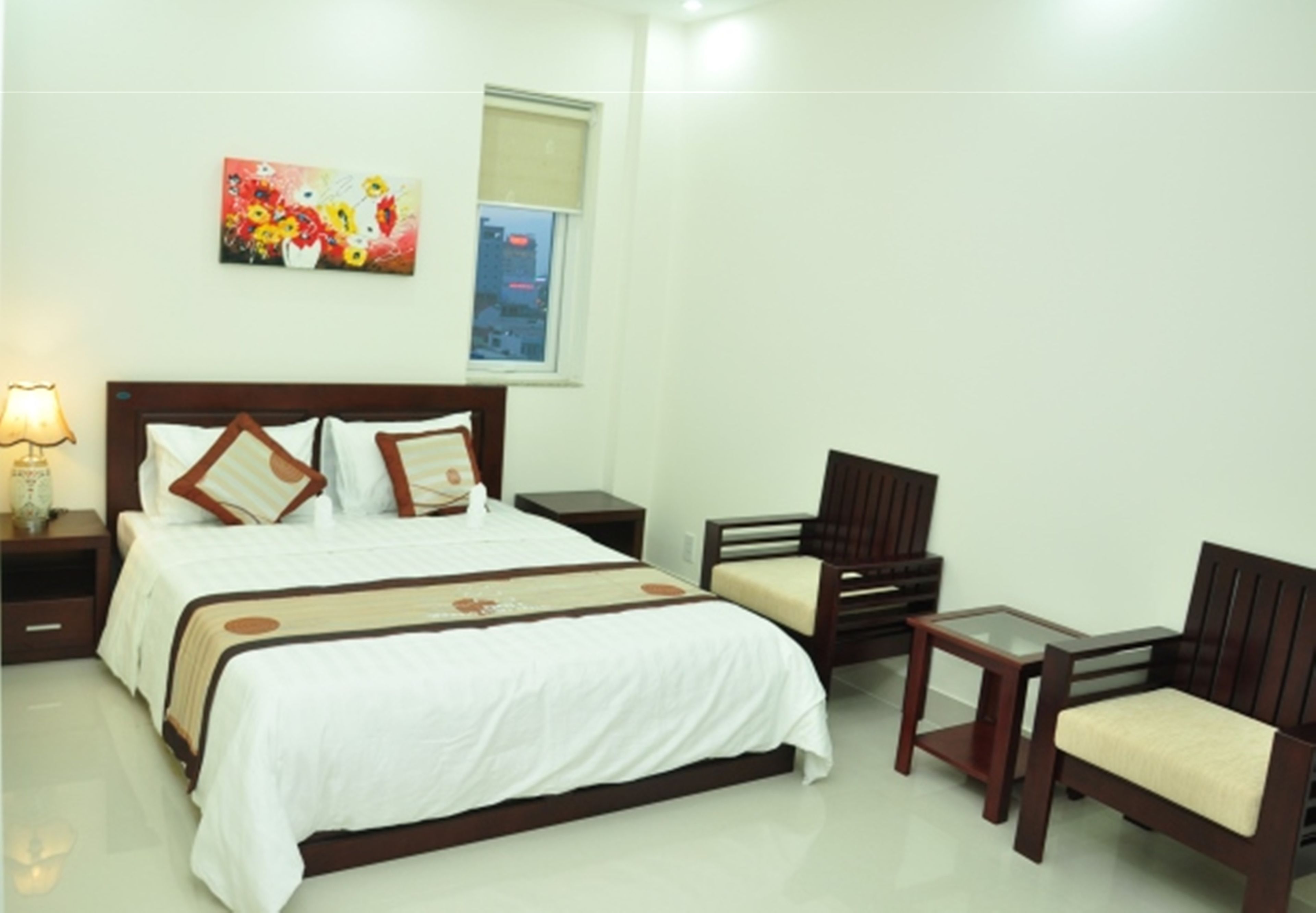 A hotel room with a bed, two wooden chairs, a small table, a nightstand, a lamp, and a painting on the wall.
