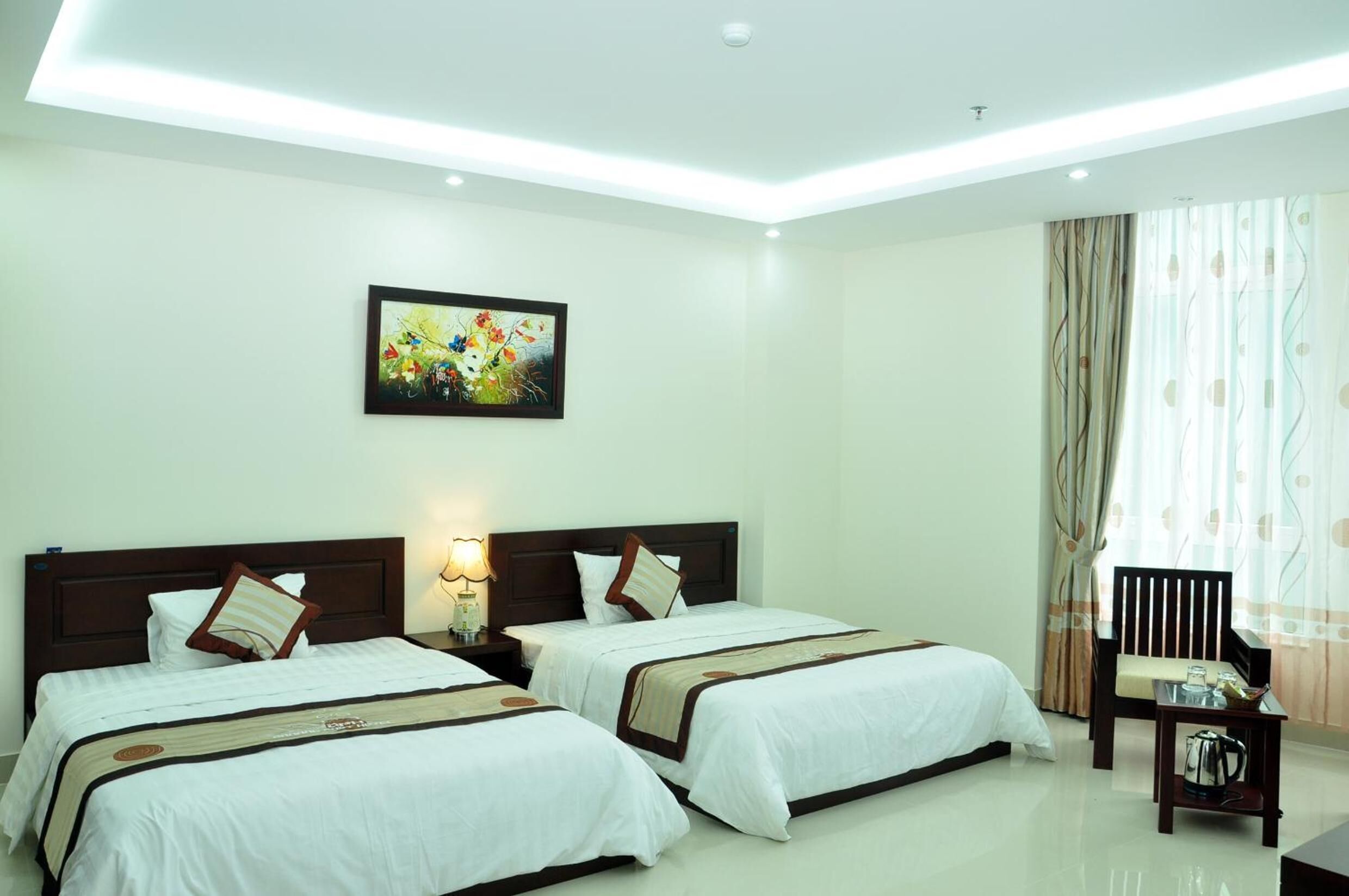 A hotel room with two beds, a nightstand, a lamp, a painting, a chair, and a table with a kettle.