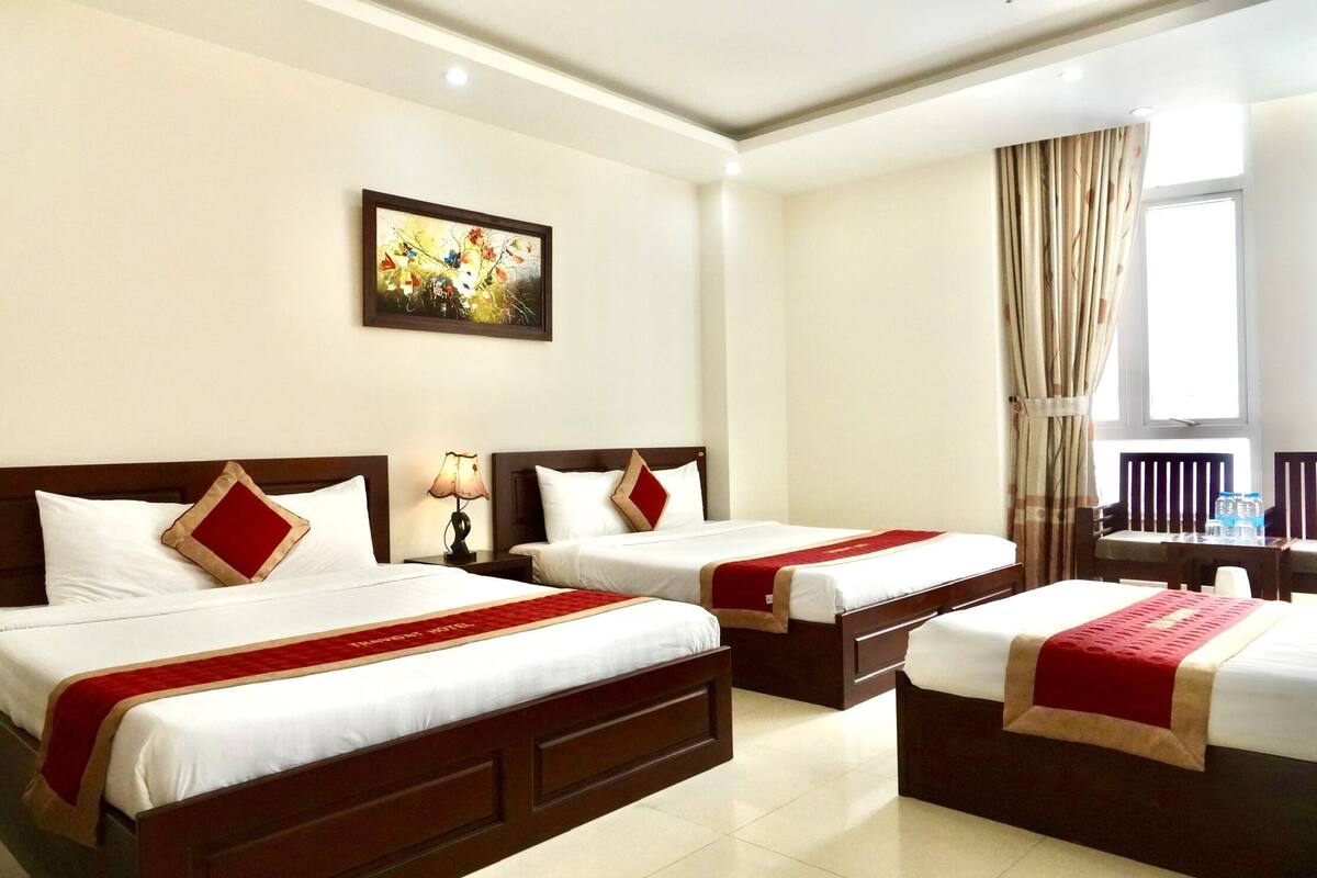 A hotel room with two beds, a painting on the wall, a window with curtains, and a desk with a chair.