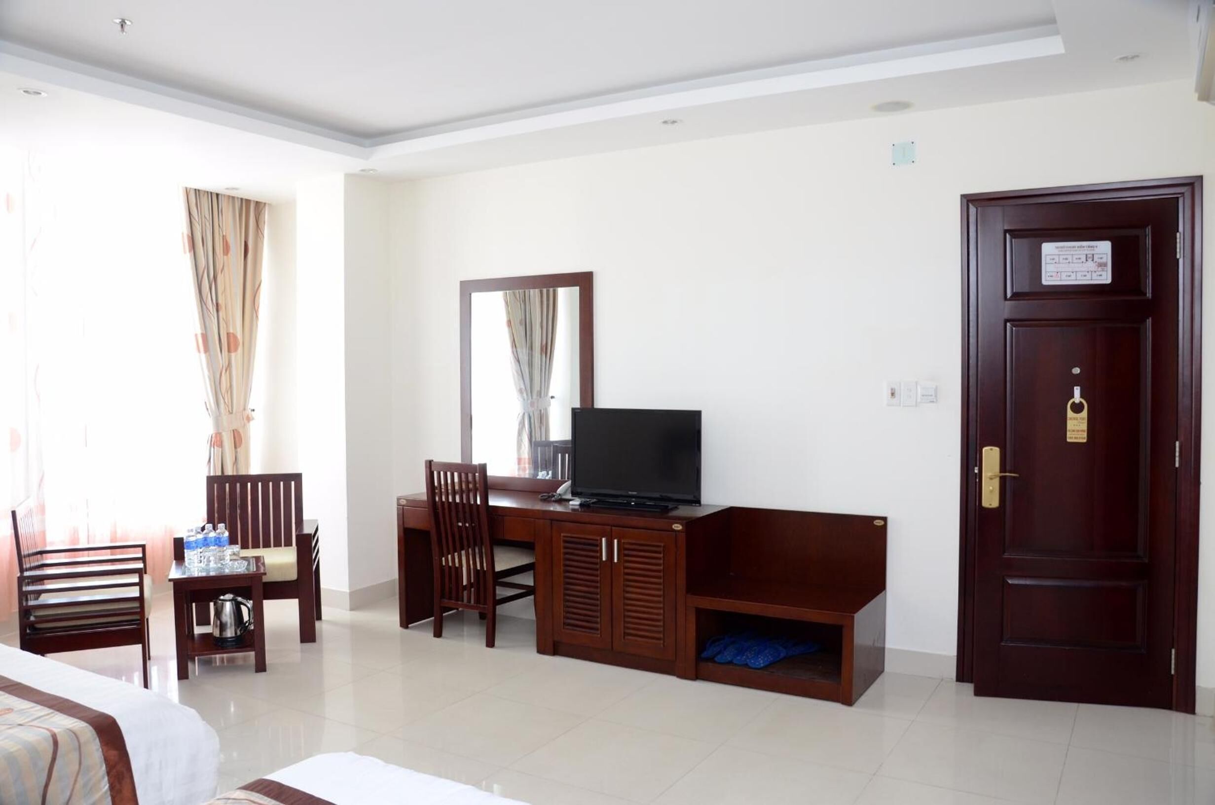 A hotel room with a bed, a desk with a television, a chair, a small table with water bottles, and a wardrobe.