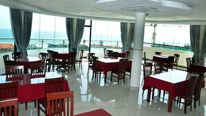 A spacious dining area with multiple tables and chairs, large windows with curtains, and a view of the sea. - Dattravi Hostel (Da Nang)