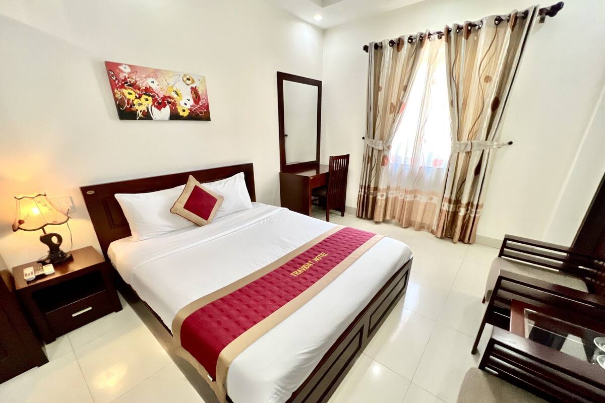 A neatly made bed with a red and white bedspread, a nightstand with a lamp, a chair, a desk, and a painting on the wall.