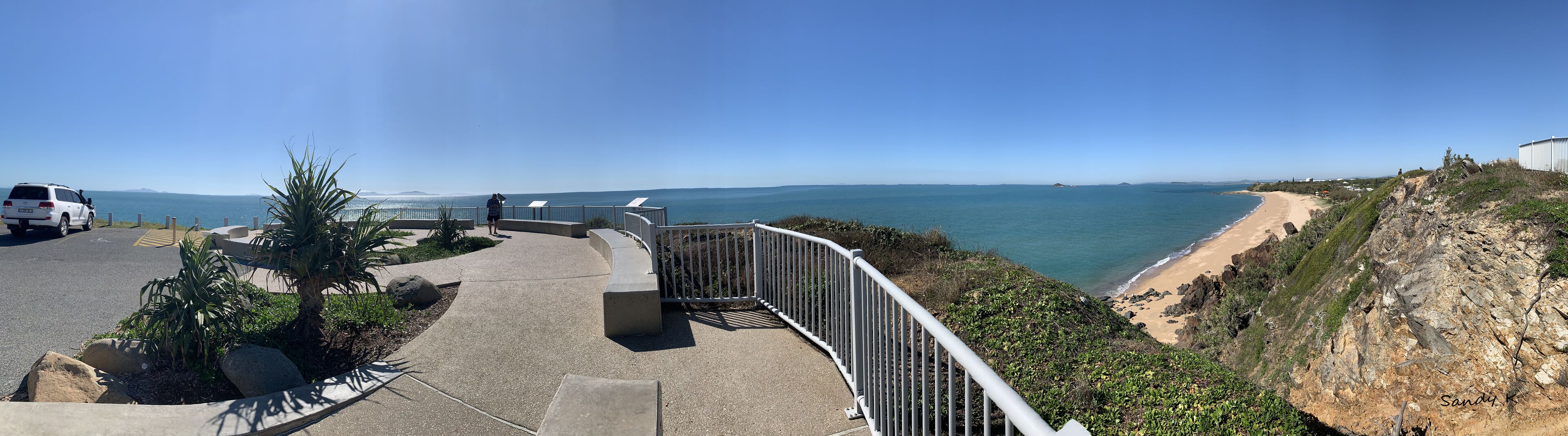 A coastal walkway with a view of the sea, a parked vehicle, and a person walking.
